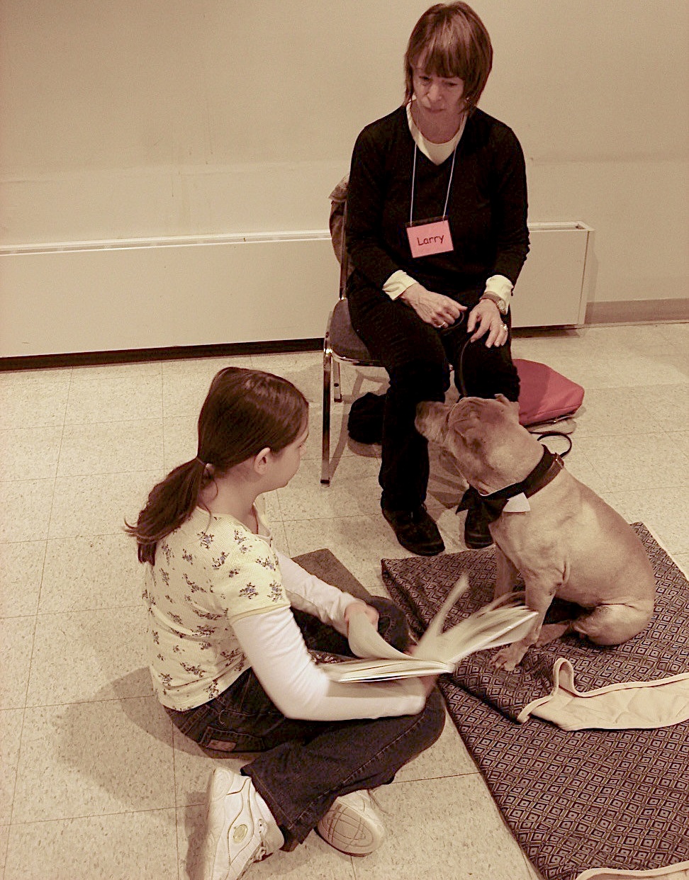 girl seated on the floor holds a book and reads to a medium brown dog; a seated woman observes from nearby