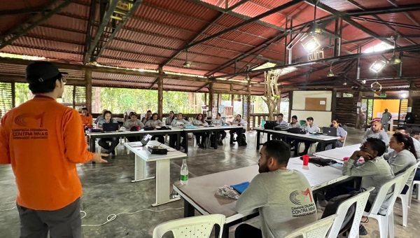  A group of people sit at tables arranged in a U shape while a man with his back to the camera addresses them.