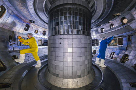 Two people, one in a yellow cleanroom suit and one in blue, work in a circular space, metallic space of a tokamak nuclear reactor.