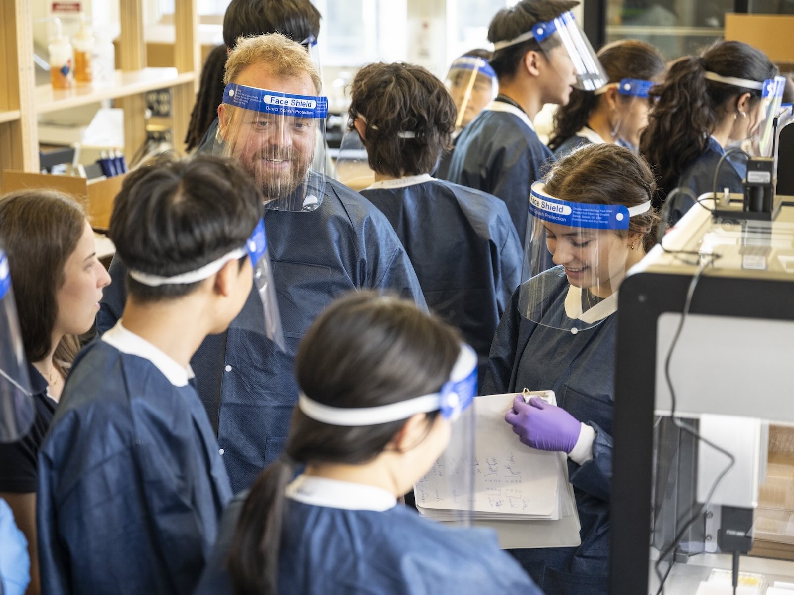 A group of people wearing protective face shields work in a lab setting.