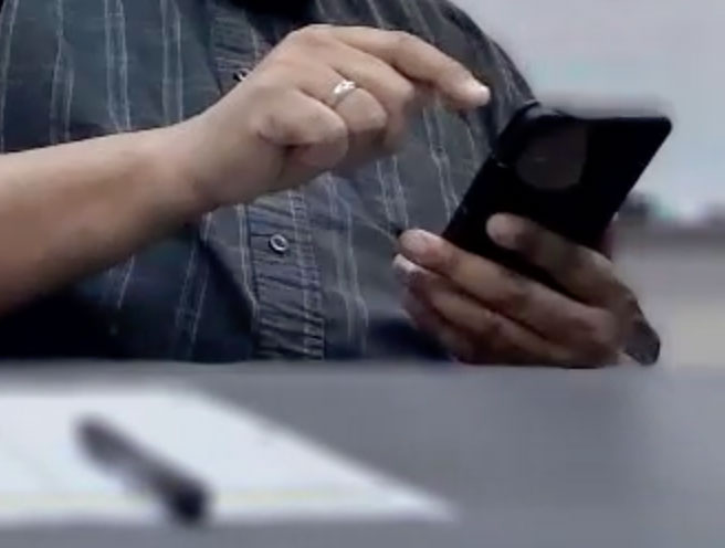  Close up of hands using a cell phone near a tabletop.