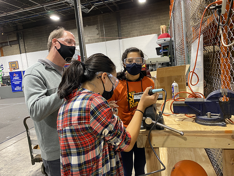 Two young girls, both with dark hair, use a soldering iron under the guidance of an adult man wearing a grey pullover with a facemask and safety glasses. 