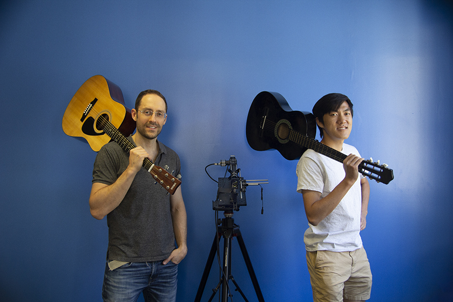 Two men separated by equipment on a tripod pose with guitars resting on their shoulders. 