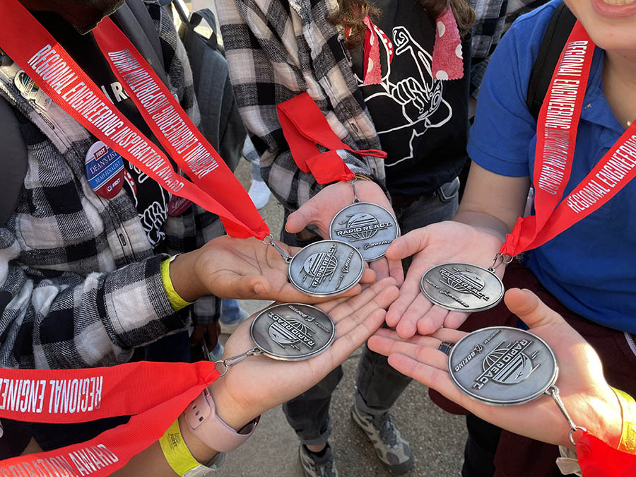 Five members of the Girls of Steel hold their medals in a circle in the middle of the photo.