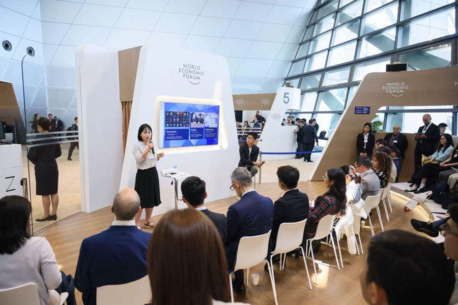  A woman holding a microphone stands in front of a modern, airy room filled with people at the World Economic Forum.