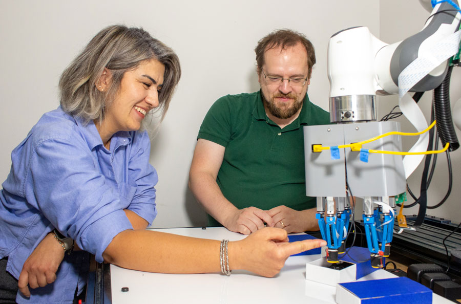  A man and woman sit at a desk with a robotic arm that has fingertip attachments. The woman interacts with those fingertips.