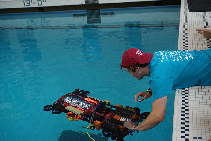 A man wearing a baseball cap backward leans over a swimming pool to launch a robotic submarine.