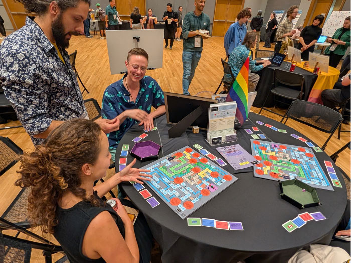  People sit around a table playing a colorful boardgame, while a man stands and looks on.