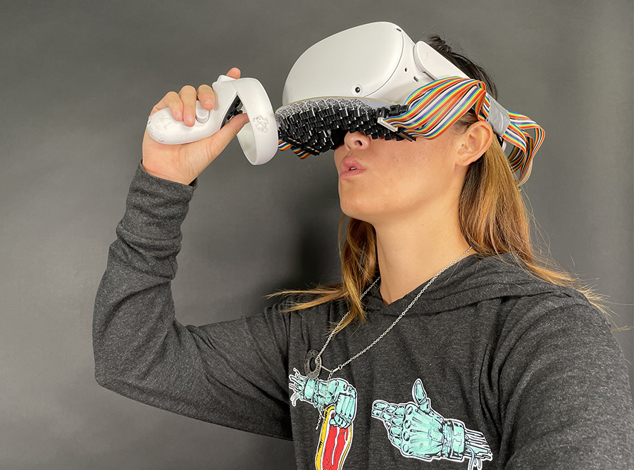 A woman wears a large goggle-like virtual reality headset outfitted with an array of sensors directly above her lips.