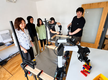 A group of people stand around a table where a robot arm manipulates a stack of colored blocks.