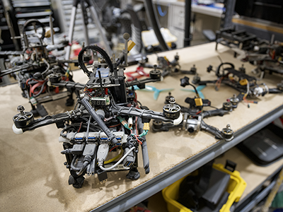  A partially assembled quadcopter drone sits on a workbench surrounded by drone frames, motors, tools, and electronic components in a workshop.