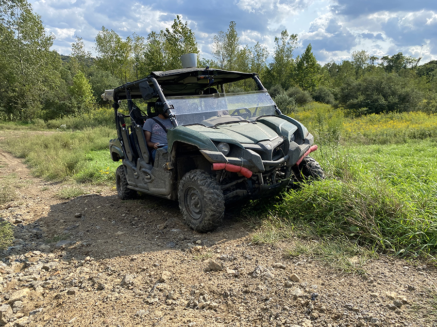 A green all-terrain vehicle outfitted with sensors sits half in the grass and half on a dirt road. Two people sit inside the ATV.