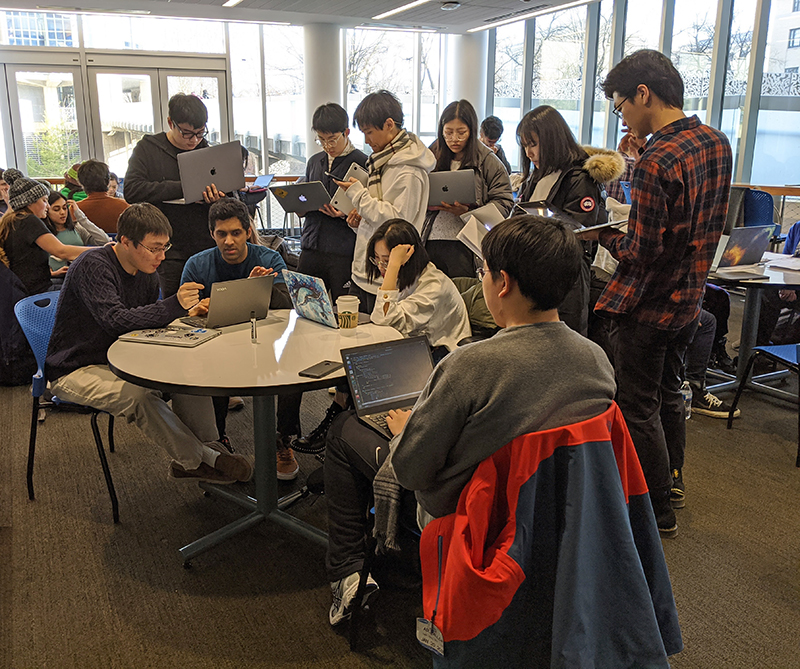 Students gather around a table and work on their laptop computers.